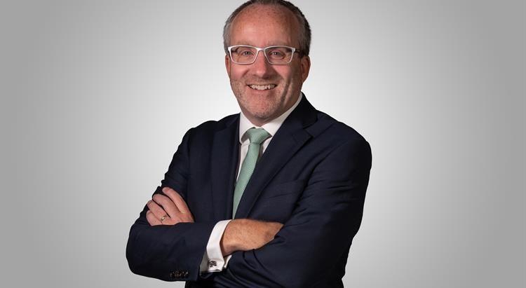 Richard Corrigan, dressed in a dark suit, white shirt, and a light green tie. He is smiling and has his arms crossed. He is wearing glasses and has short dark grey hair. He is in front of a plain background.