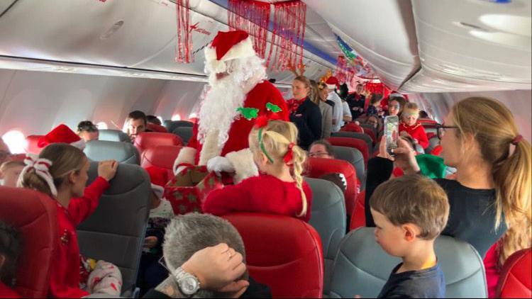 A man dressed as Santa walks down the aisle of a passenger plane handing out presents to children in Christmas outfits.