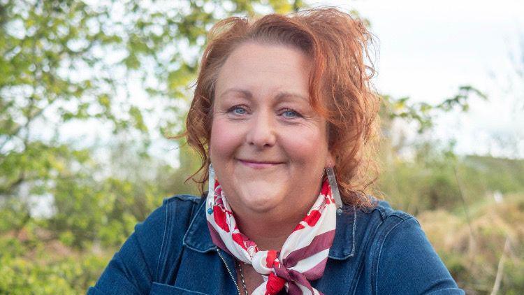 Janet smiling at the camera. Janet has red hair and blue eyes. She is wearing a denim blue top and a white and red scarf around her neck. She is outside, there are trees with green leaves in the background.