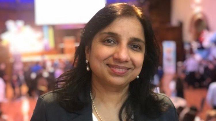 Dr Sumeeta Gupta has long black hair and is smiling. She is wearing a black blazer and white top with a light-coloured necklace.