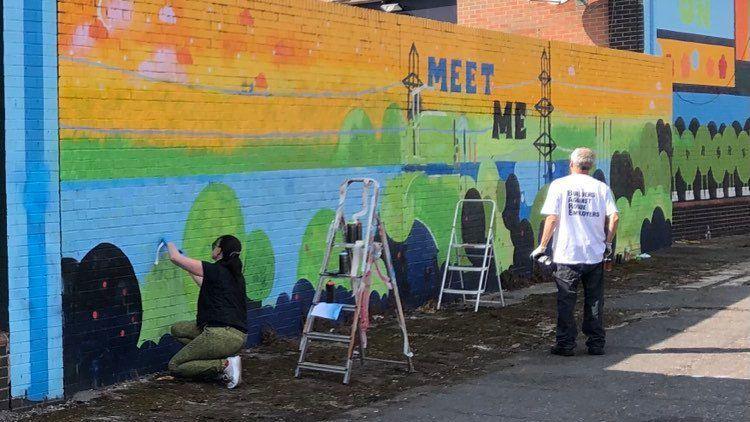 Residents taking off paint - a woman in the foreground on her knees scrubbing at a wall with two stepladders near the mural which says meet me.