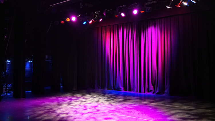 A theatre stage. Purple strobe light falls on an empty stage in front of a closed curtain