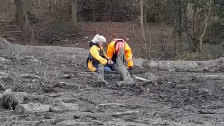 A woman with grey hair, wearing a yellow coat, being helped out of some mud by a man in rescue gear. There is a lot of dark-grey mud in the foreground.