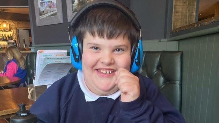 Thomas, 12, who has black hair and is wearing blue ear defenders and a blue jumper, smiles at the camera while sitting on a table at a restaurant