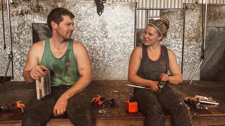 Aaron Magee with short brown hair is sitting on a stage to the left. He is wearing a dirty green vest top which is covered in sheep dirt and sweat. He is sitting beside a girl with blonde hair in a black vest top, she also has dirt on her. She is holding shearing equipment. There is also equipment beside them. There are silver metal panels behind them.