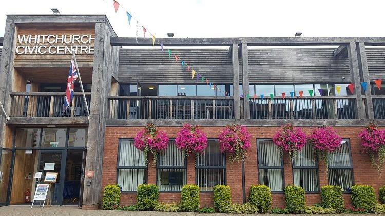 The front of a two-storey building. The first floor is made of red brick and has hedges and purple hanging flowers in front of it. The second storey is mostly clad in timber. A sign on the second storey says Whitchurch Civic Centre
