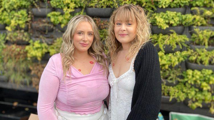 Ellie (left) wears a pink long-sleeved top. She has long blonde wavy hair and fake kiss tattoos on he chest. She stands with a friend who has blonde shoulder-length blonde hair and a full fringe. She wears a white to and a black cardigan