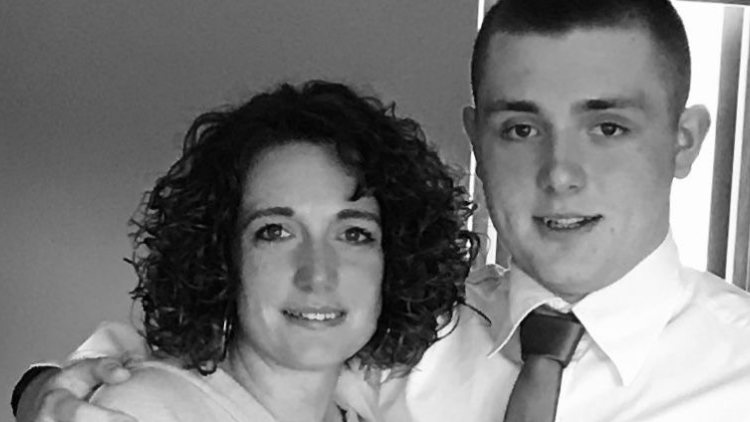 A black and white image of woman with dark curly hair and a young man, her son, holds her round the shoulder in a tie and white shirt