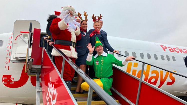 A man dressed as Santa and a man dressed as an elf join cabin crew on the steps leading up to a white Jet2 plane with red writing on its side