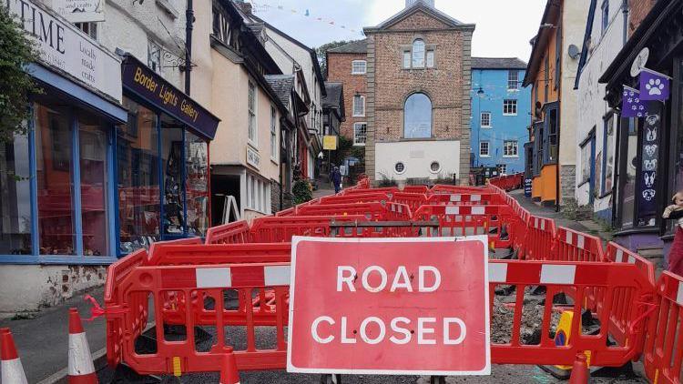 A red "road closed" sign in front of dozens of red traffic barriers, which have been placed up a street. There are shops either side of the road, and a building at the top of the road with a large rounded window 