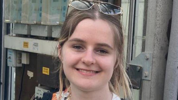 A head and shoulders picture of a young blonde girl wearing sunglasses and smiling at the camera.