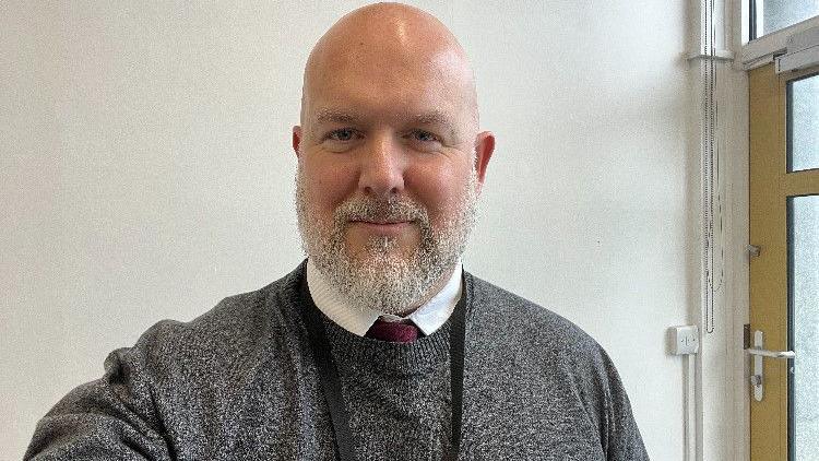 A head and shoulders shot of Chris who is a bald middle-aged man with a grey beard. He's wearing a grey jumper and a tie.