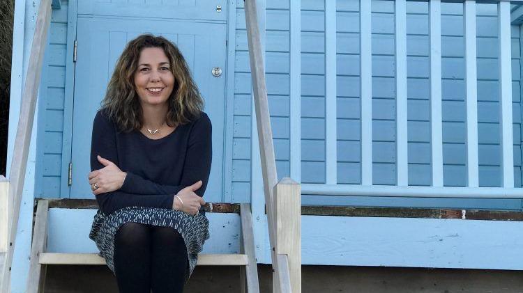 Hetti Simpson is sitting on the steps of a blue beach hut, smiling with her arms folded. She's wearing a dark blue top and patterned blue and white skirt.