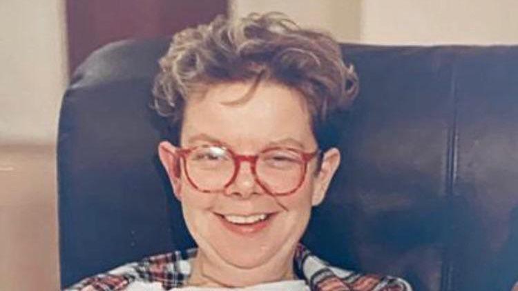 A photo from the 80s of a woman in a leather arm chair wearing round red glasses and smiling