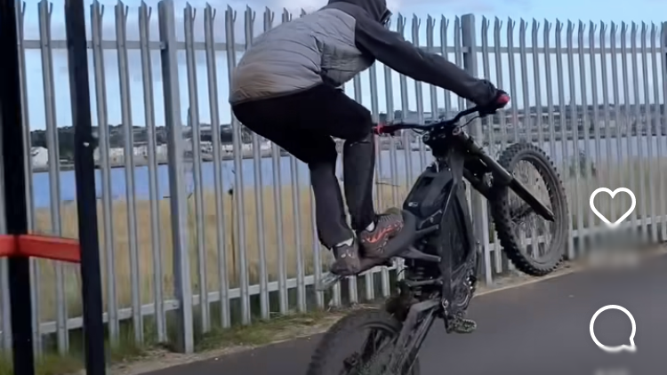 An e-bike rider with hood up and standing on the saddle doing a wheelie, with front wheel high in the air on what appears to be a path next to a fence