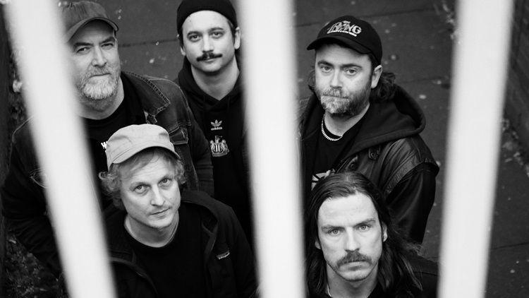 An aerial photograph of the band grouped together looking above them into the camera. There are bars in front of them. They are standing on a path and are all wearing black. The man standing in the middle  is wearing a jumper with the Newcastle United club crest in the centre.