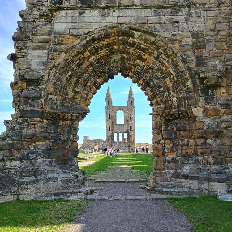 A view of the ruined cathedral in St Andrews through a stone brick archway. A pathway links the two and grass surrounds the path. There's a bright blue sky and tourists wander in the background.