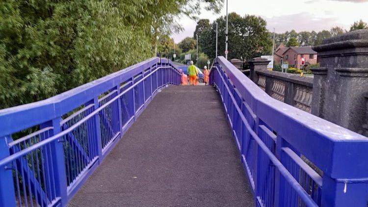 A temporary footbridge with blue plastic railings and workmen seen in the distance. It runs parallel to a road bridge, of which stone barriers can be seen to the right.