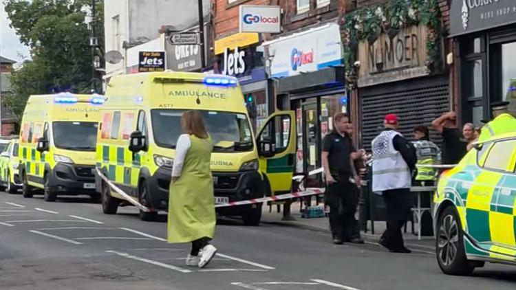 Image shows a street closed off, with paramedics and their vehicles standing on the pavement with members of the public. Red and white incident barrier tape can be seen around one of the ambulances.