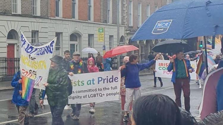 Pride marchers holding signs about the Presbyterian church supporting LGBT communities. It is raining and many people have an umbrella up. 