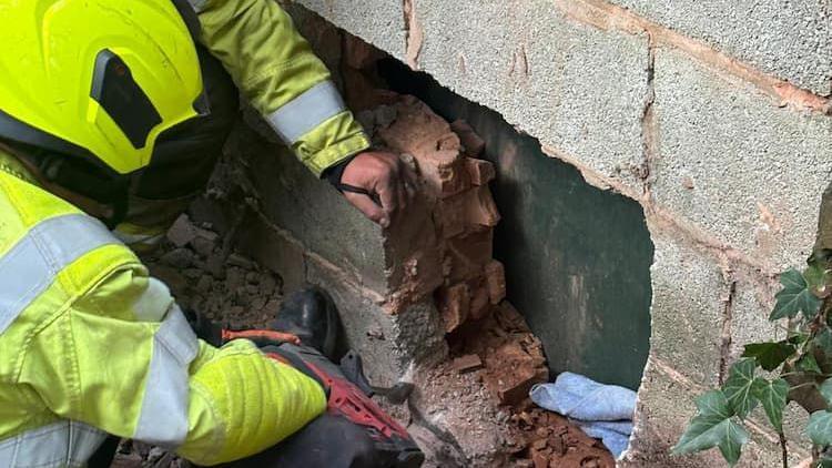 A firefighter is wearing a yellow high-vis jacket and helmet. He is drilling through a wall in an attempt to reach a kitten.