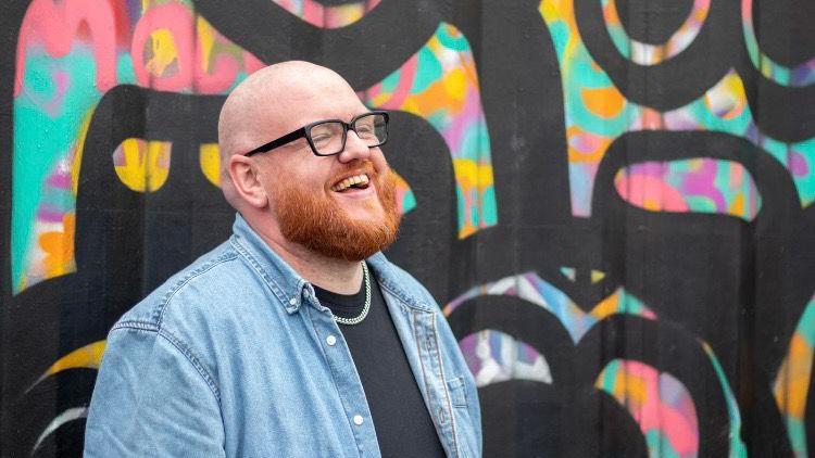 A man with a ginger beard throws his head back and laughs. He has black rimmed glasses, a denim shirt and a silver chain. 