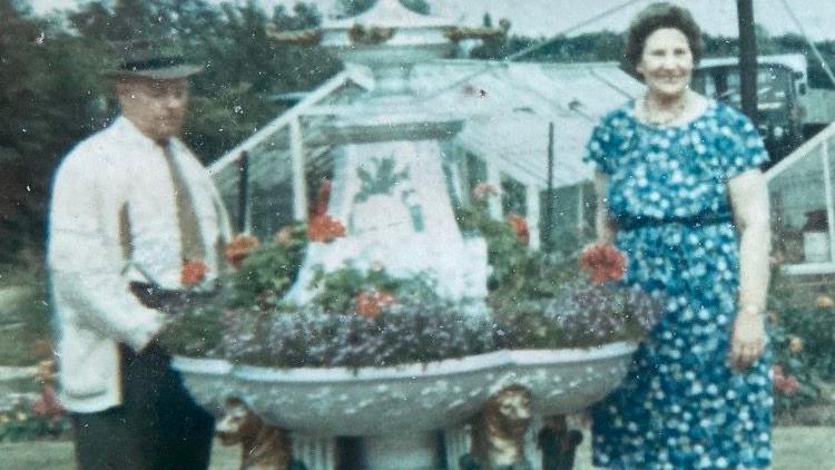 A man with a pork pie hat and white cardigan over a white shirt and brown tie next to a large drinking fountain that has had various blooms planted in it. The man's wife is on the right, she has dark permed hair and a floral dress on with a necklace around her neck. A greenhouse is visible behind them