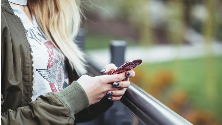 A close-up of a woman with long blonde hair wearing a green jacket and white top texts on a mobile phone, positioned next to a barrier with a grass area to her left. Her face cannot be seen.