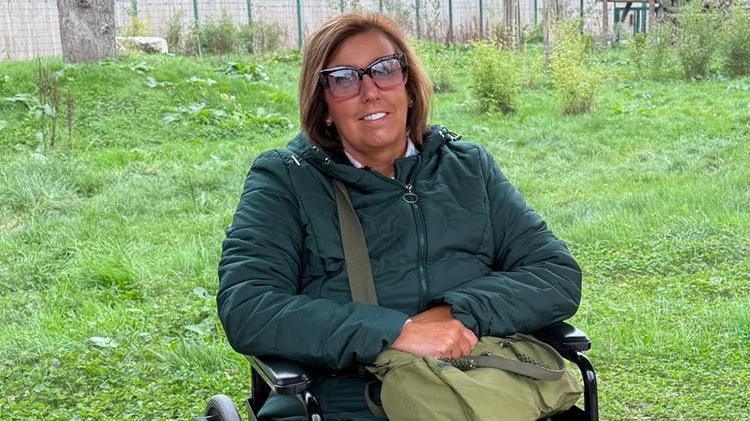 Emma Martin with brown hair in a bob wearing a dark green coat and holding a light green bag on her lap with a grass area behind her fenced off with a green fence. She is using a wheelchair. She is smiling. 