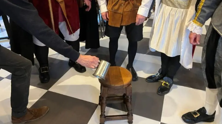 Five people stand around a wooden stool, with one person leaning into the circle to pour ale on the stool from a tin cup.