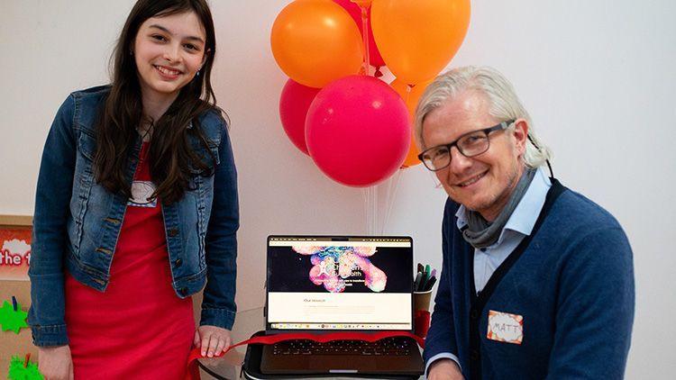 Matt Zilbauer is sitting at a table in front of a laptop. He has white medium length hair and is wearing glasses, a blue shirt and blue cardigan. He has a sticker with his name on stuck to him. Standing next to the table is Holly. She has long dark hair and is wearing a red dress and denim jacket.