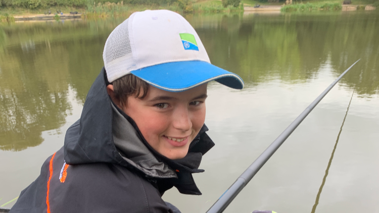 Charlie smiles at the camera, wearing a white and blue cap and a black coat with his fish rod hanging over the lake in the background.