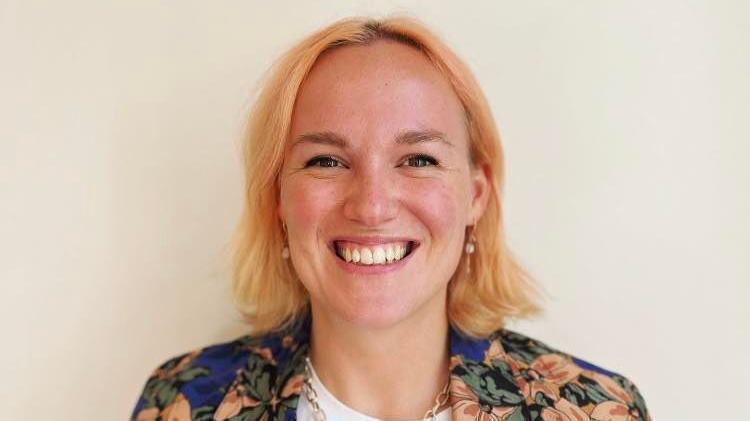 Picture of Dr Hester Hockin-Boyers, smiling at the camera widely. She has pale pink/orange short hair and wears a brightly blue and orange flowered blazer. She has hazel eyes and wears gold earrings and a gold necklace. 