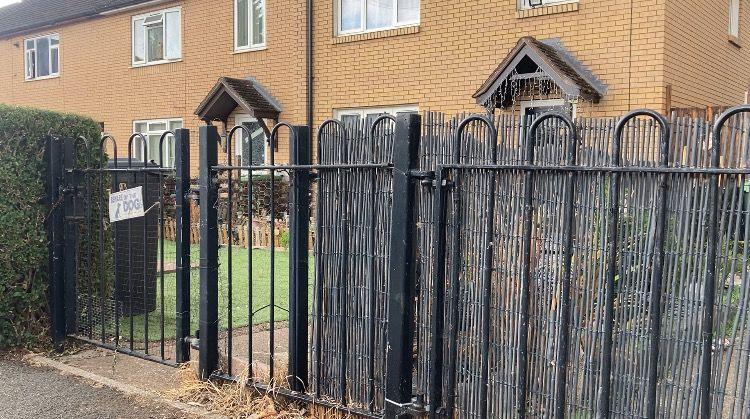 A row of beige brick houses. The nearest house sits behind a black metal gate and fence. There is a path running to the house from the gate and some grass to one side. There is a sign on the gate that says 'beware of the dog'.