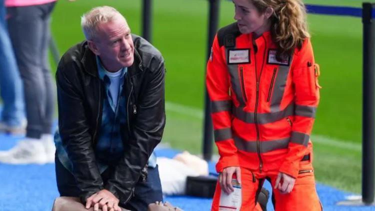 DJ Fatboy Slim wearing a black leather jacket on his knees delilvering CPR to a dummy with an air ambulance professional next to him. She is wearing orange overalls 