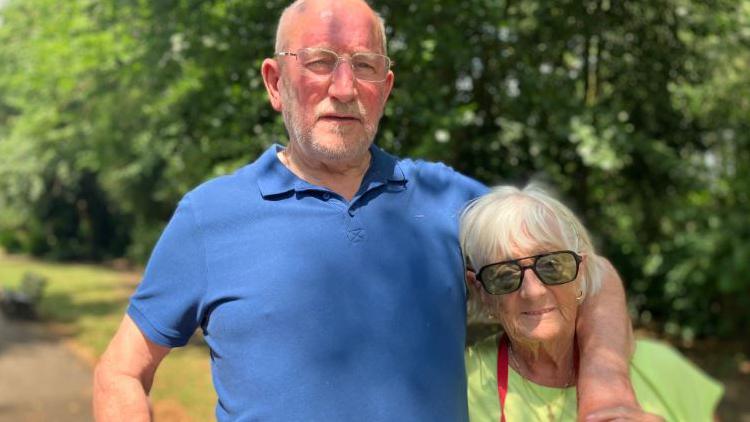 A man in a blue t-shirt wearing glasses with a slight grey beard, with his arm around his wife who has short white hair and is wearing sunglasses and a yellow top.