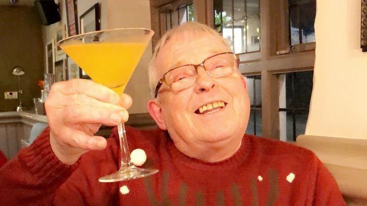 A grey-haired man with glasses smiling and holding up a yellow cocktail