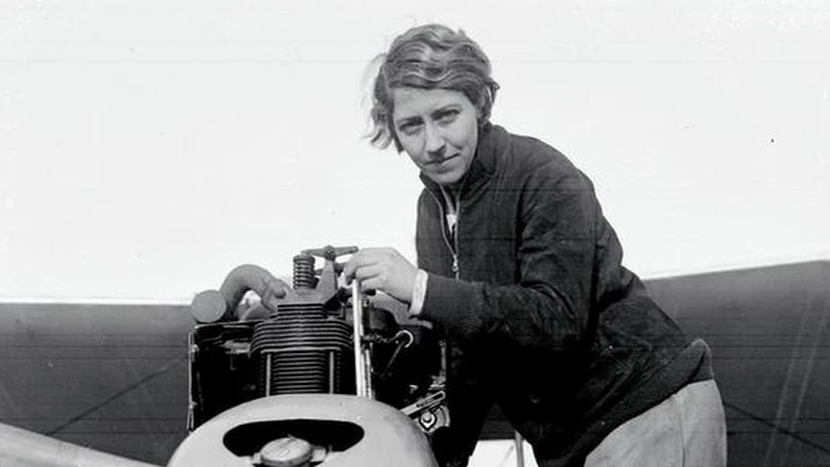 Black and white photograph of Amy Johnson dressed in flying gear in the cockpit of an aircraft. She is looking towards the camera and smiling, with her goggles pulled up on her forehead.