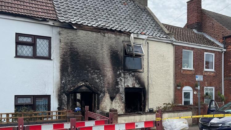 The house, painted in cream colour, has black smoke and fire damage above the ground floor door and window, with damage caused to the fascia boards and upper floor window. Red and white temporary fencing creates an alternative walkway for pedestrians owing to the pavement being excavated for utility access for safety reasons.
