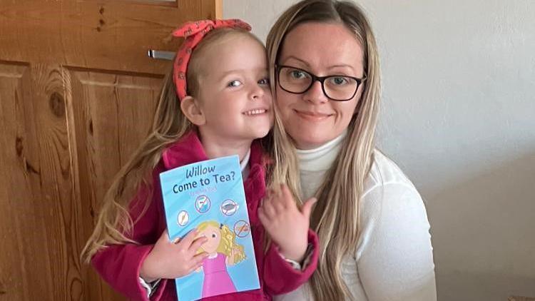 Willow wearing a pink coat stands next to her mum, holding a copy of the book.