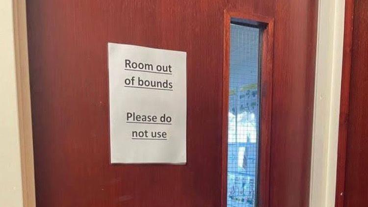 A brown door with a long skinny window to the left built into it. There is a piece of paper with "room out of bounds, please do not use" written on it. 