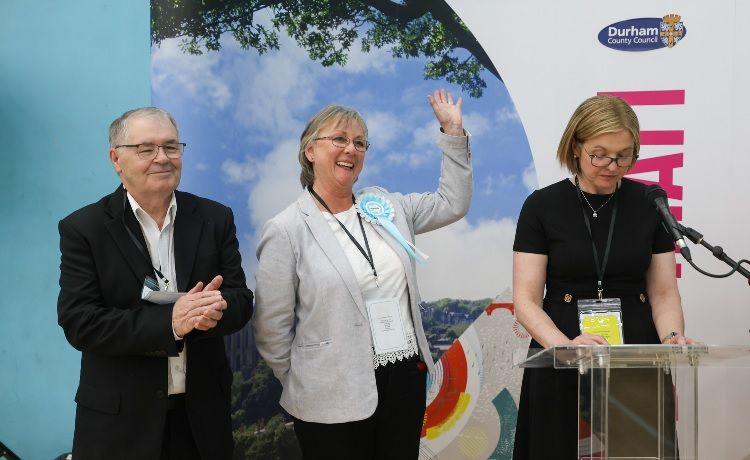 John Bailey, who has grey hair and glasses, wears a black suit and is clapping. His is standing next to Louise Taylor who is waving. She has short grey hair and wearing a light coloured top. She is smiling. They are next to a woman who is reading out election results.