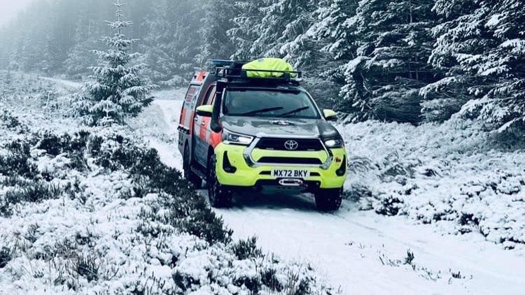 A mountain rescue vehicle goes up a snowy mountain road with snow covered trees on either side.