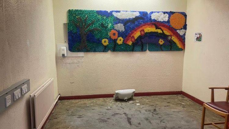 An empty room with grey walls and grey flooring. There is an art piece hanging on the back wall with a tree, flowers, a rainbow and the run on it. There is a broken sink on the floor beside the back wall and a radiator hanging on the left wall. 