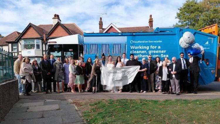 A blue bin lorry in a residential road with wedding guests standing next to it.  One is holding balloons.  There are residential homes in the background and a garden at the front of the picture.