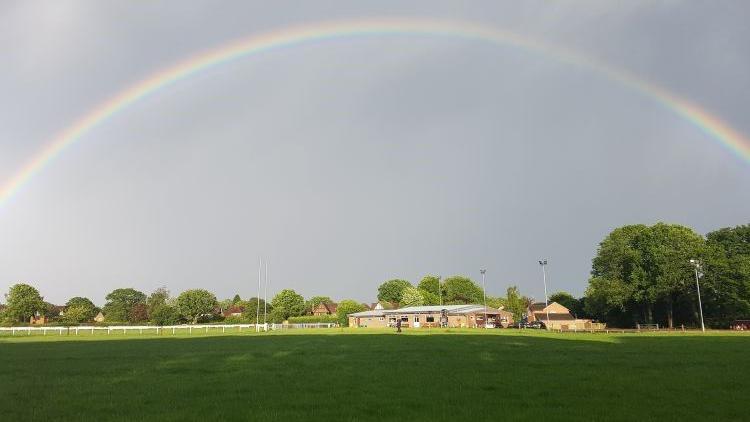A rainbow stretches perfectly across a grey-blue sky. Below is a large green field with a rugby goal in the distance and floodlights. A brick built single storey club house is at the centre of the picture, with trees around the edge. The field is mostly in shade with sun on the buildings and trees.