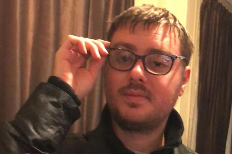 The photo shows a man looking at the camera with a straight face. He's wearing black glasses, and is touching them with his hand. He's wearing a black jacket. 