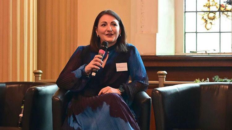Ema Cubitt is speaking in to a microphone. She is seated on a leather chair. She is wearing a blue and black patterned dress. 