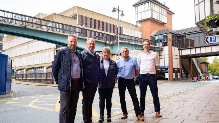 Four men and one woman standing in front of a footbridge and a car park.

