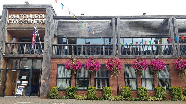 The entrance to Whitchurch Civic Centre. The building's ground floor is red brick while the exterior of the floor above is wooden. There are six pink hanging baskets, a union jack and bunting at the front of the building. 